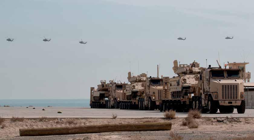 UH-60 Black Hawk helicopters fly over armored vehicles loaded on transport trucks during Army Day 2018, February 9, 2018, Kuwait Naval Base, Kuwait (U.S.