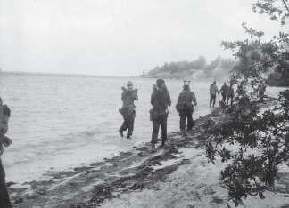 Photographed on April 19, Cuban troops carry mortar equipment along a beach; the SS Houston is seen still ablaze in the distance, which may place these men on the western shore of the Bay of Pigs,