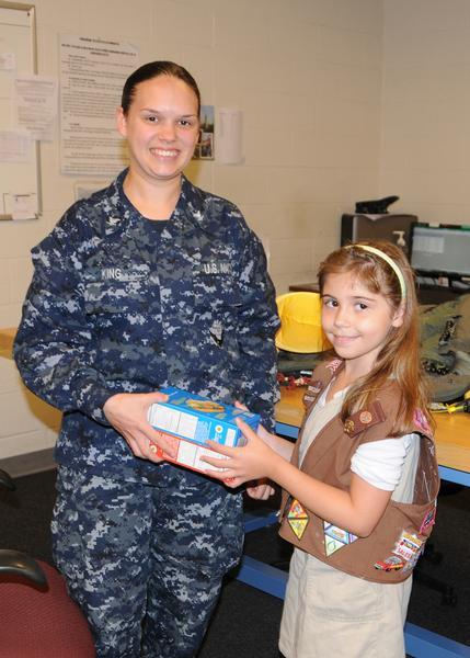 PR3 Coty Kissinger (from left) holds two boxes of Girl Scout cookies presented by Lyric Etienne who sits on the lap of PR2 Nyomi Ellis during a visit to the Paraloft in Hangar 1000 on Naval Air